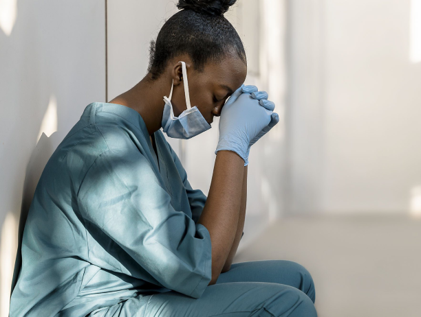 A healthcare worker in scrubs and gloves sits with head bowed, deep in thought.